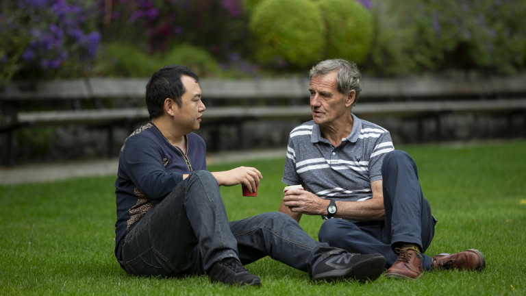 Two men sitting on the grass in a park with coffee cups, talking. 